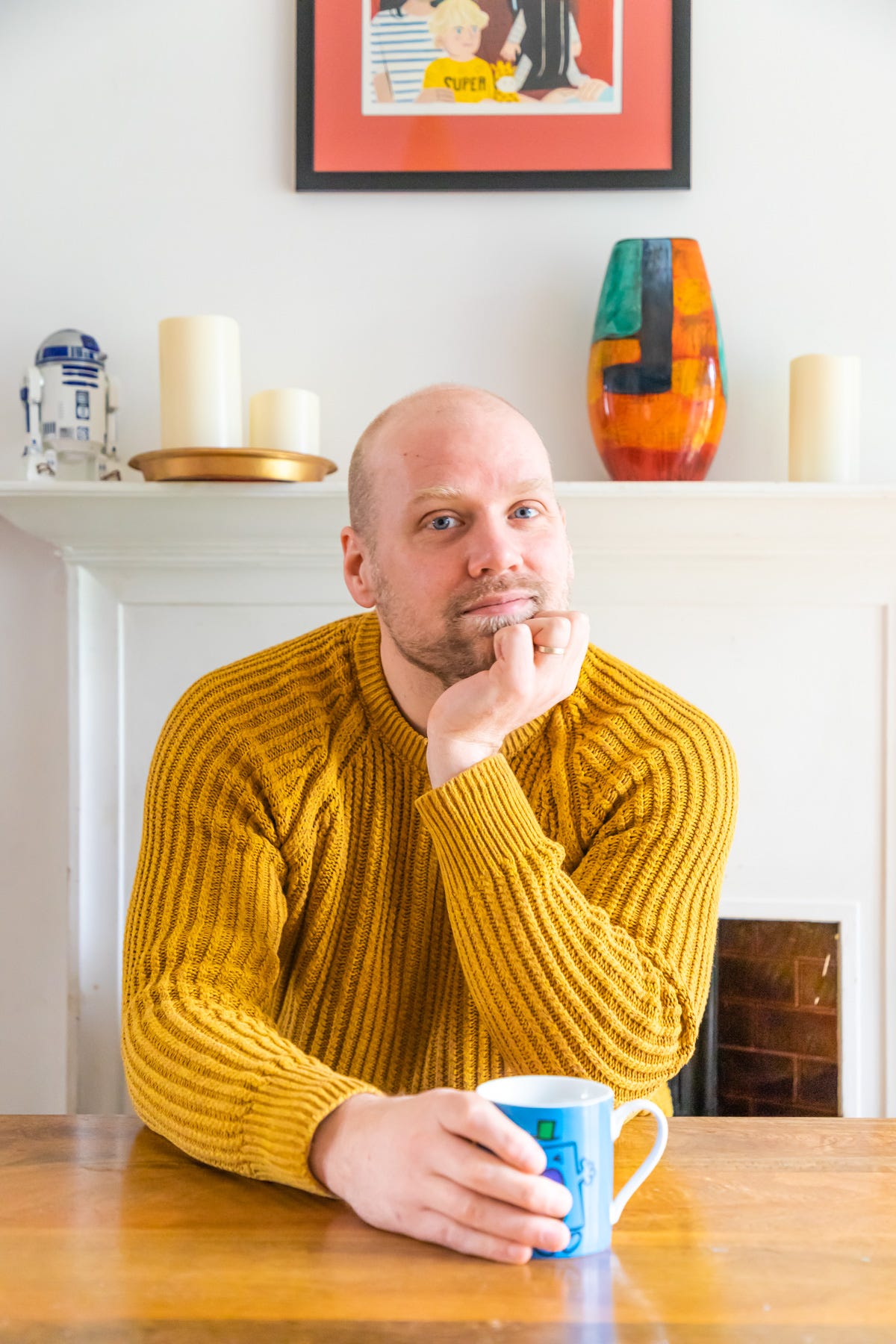 Stu - a white man - sits at a table wearing a mustard jumper clutching a Mr Man mug.