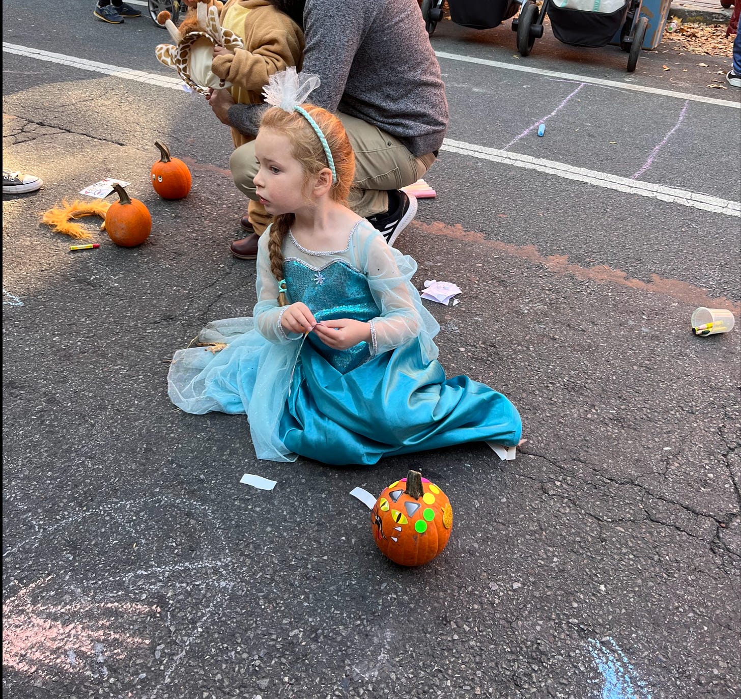 M, who is five years old, sitting in the middle of the street, wearing an Elsa costume and putting stickers on a small pumpkin.
