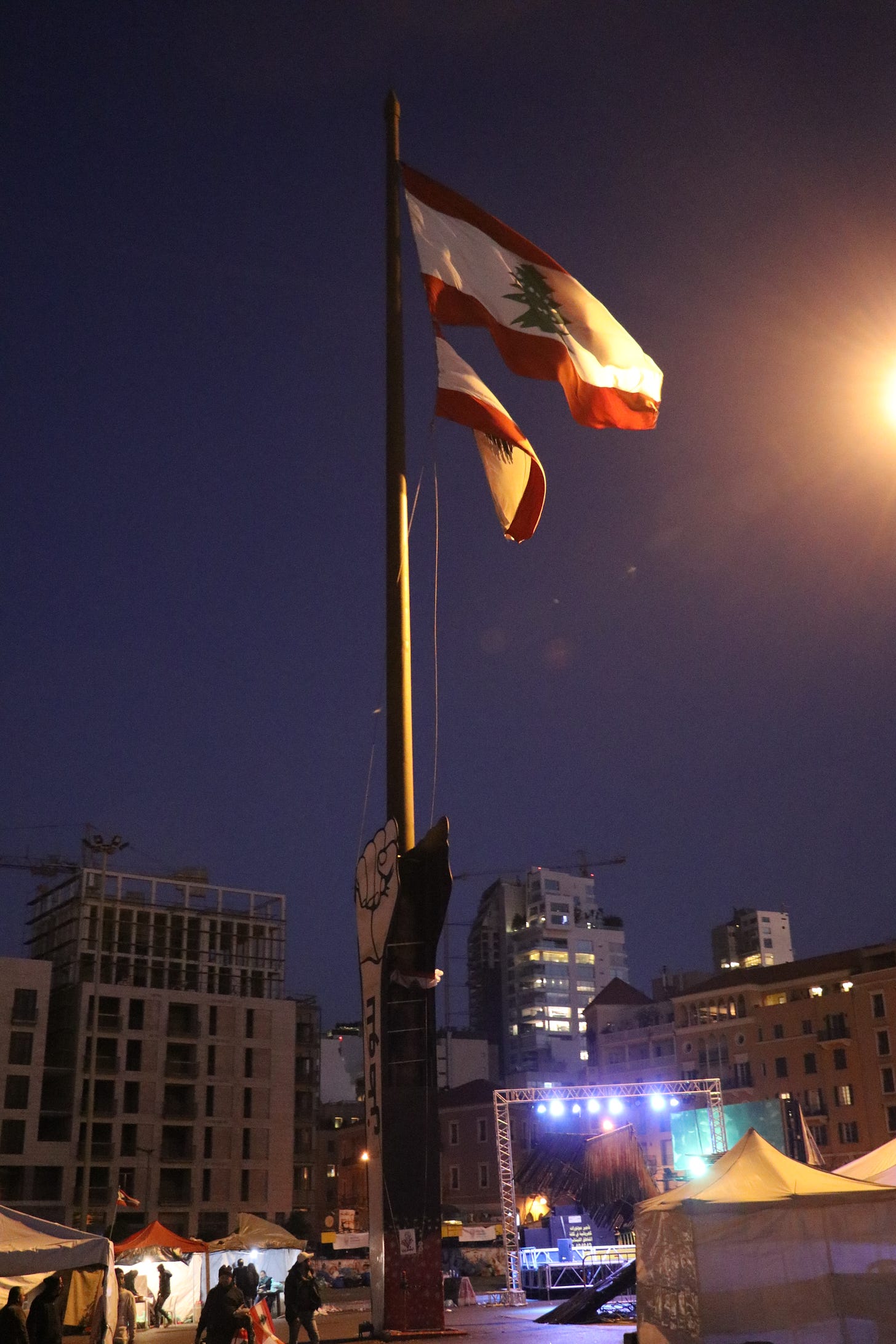 A tall monument in the shape of a raised fist holds a flagpole, atop which sit two large Lebanese flags. Tents and a stage surround the monument.