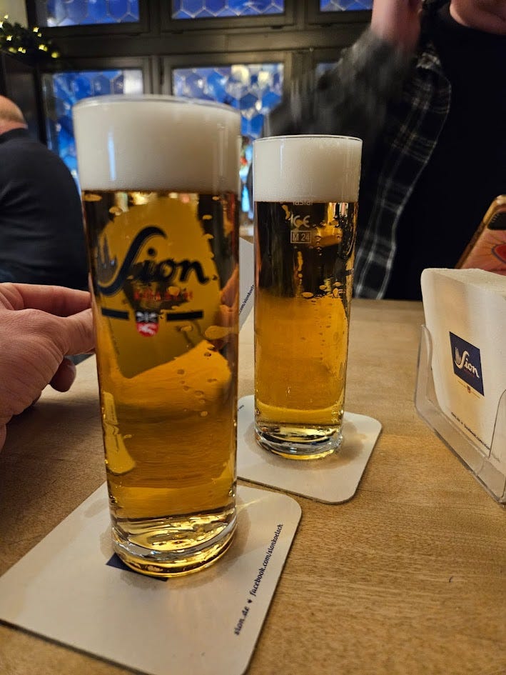 A blurry photo of two glasses of Kolsch beer with the Sion logo printed on in black. Behind you can see fairy lights and a panelled window.