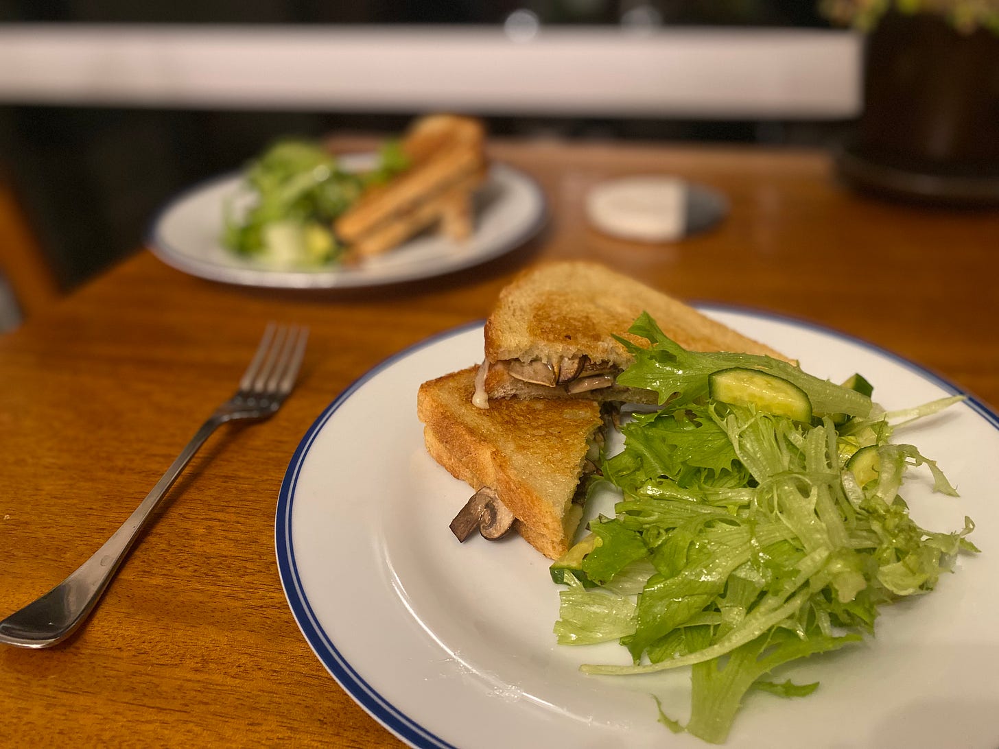 Two white plates with blue rims, each with a green salad and two halves of a diagonally sliced mushroom grilled cheese. A fork is next to the plate on the table.