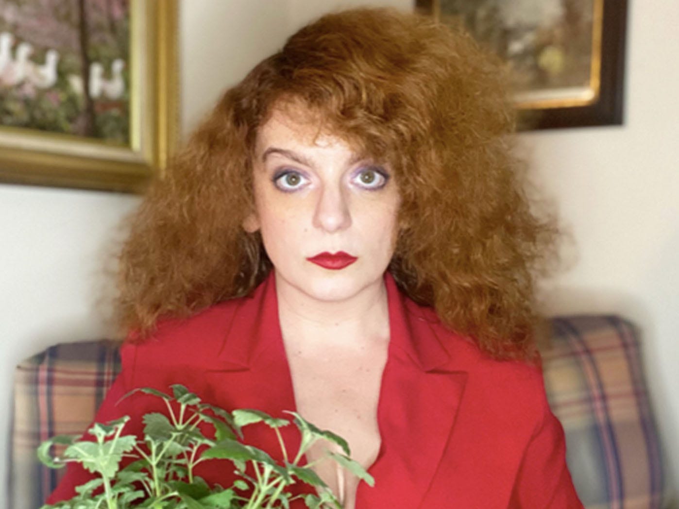 A photo of writer and performer Sara Benincasa looking quite serious as she dons a stylin' red blazer and holds a houseplant.