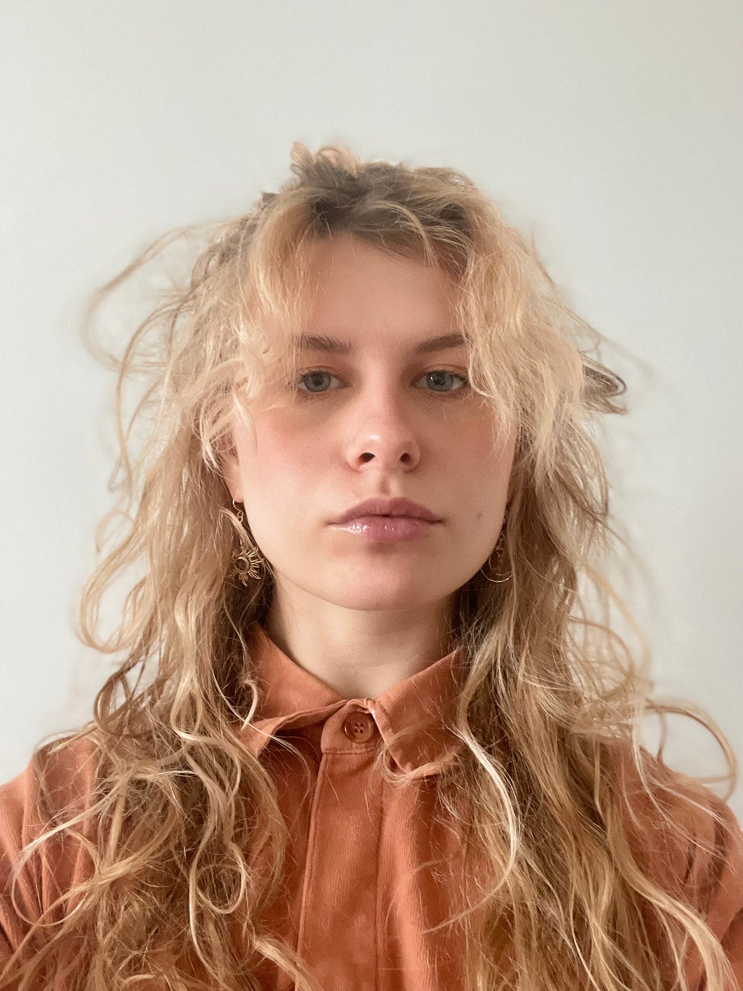 an ID style photo of a white woman with wavy hair, pale orange jumper and gold sun/moon earrings she got from her partner for Christmas, a solemn grumps look