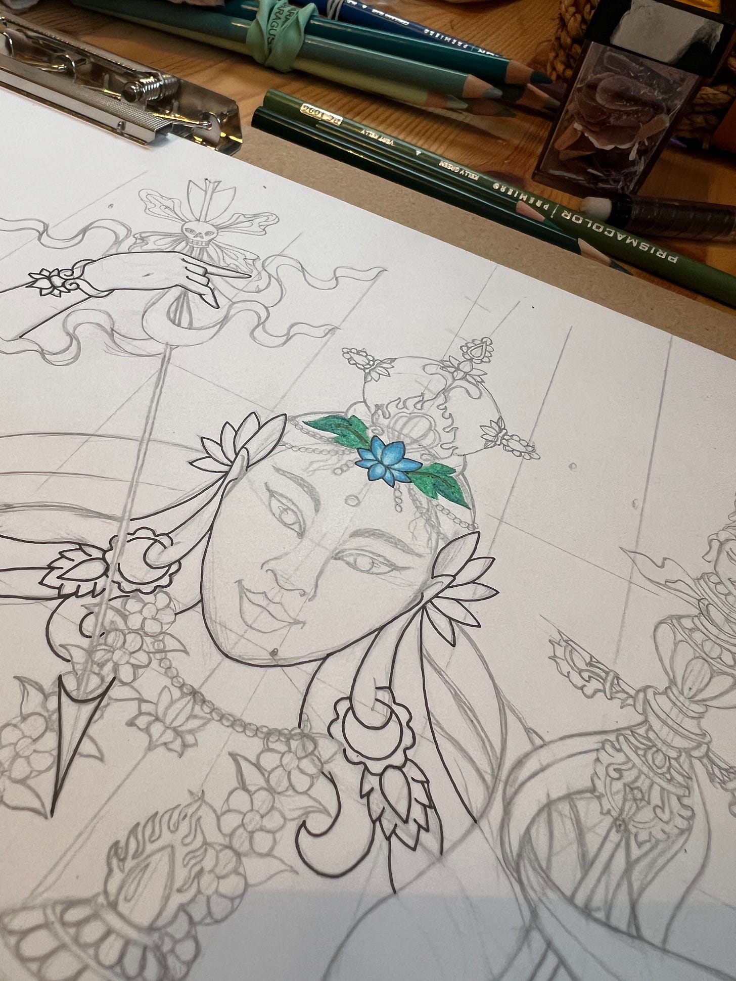A progress photo I took of Princess Mandarava the day I started to add colour to the image. The photo captures the figure from the top half, their face partially outlined in fine line black ink. They have a lotus flower in the middle of their forehead, which has been coloured in blue, with green leaves either side of it. 