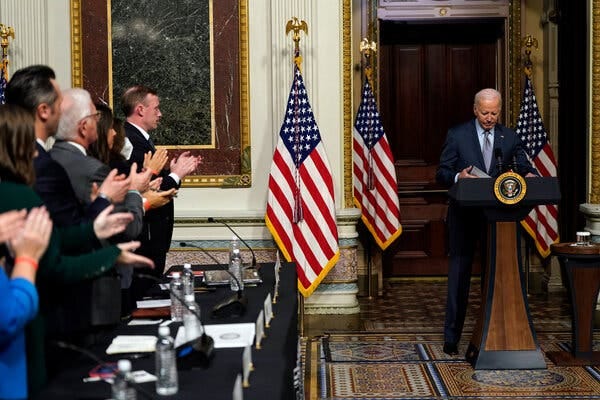 A row of people behind a black table stand up and clap for President Biden, who is reading from behind a lectern in an ornate hall.