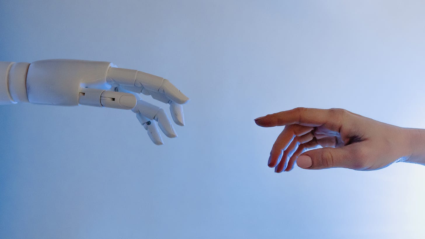 Image showing a white robot hand on the left, reaching towards the middle of the image, and a medium-toned human hand reaching in towards the middle from the right side.