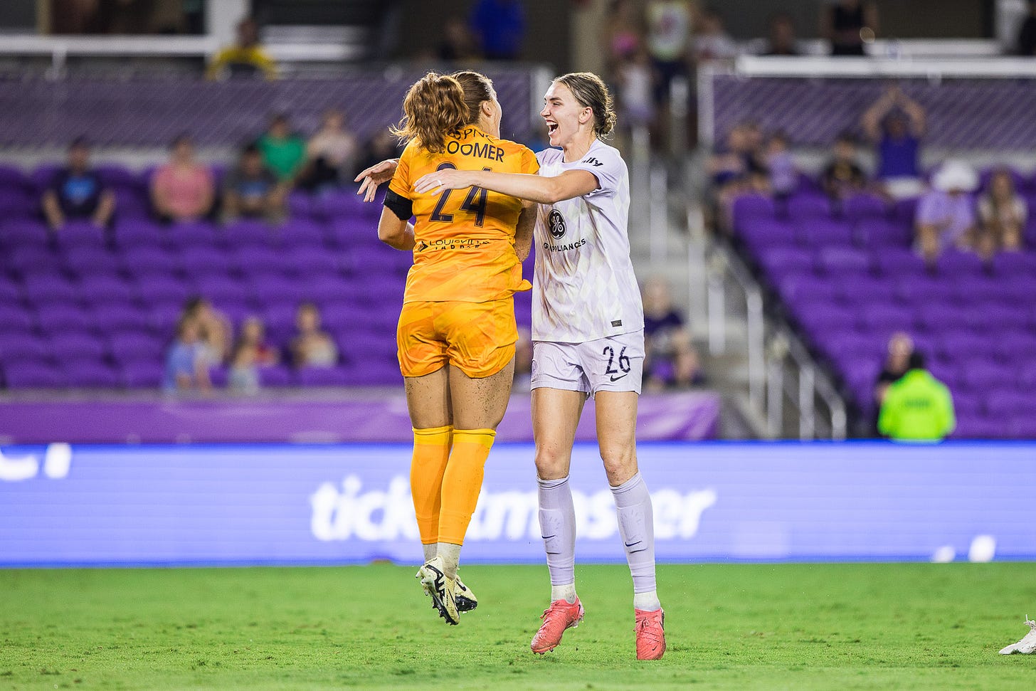 Woman in yellow gk kit with back turned jumps vertically in arms of woman in whit field kit facing forward