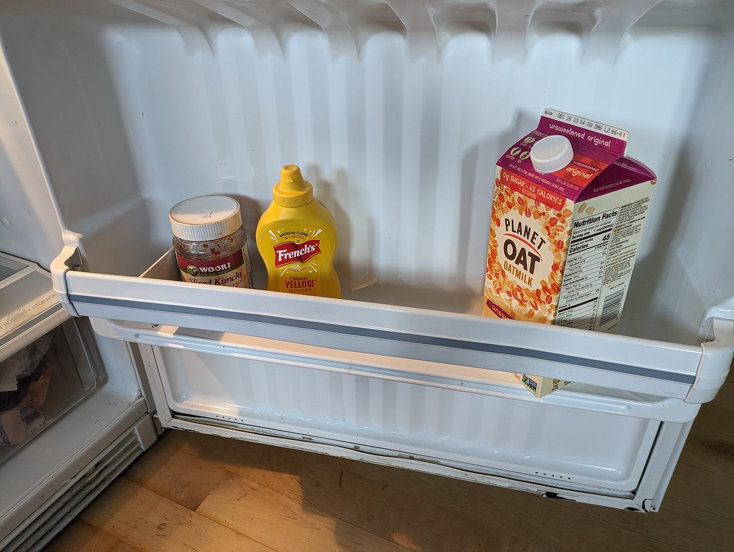 a shelf in a refrigerator door containing only three items, a half gallon of oat milk, a bottle of mustard, and a jar of Kimchi