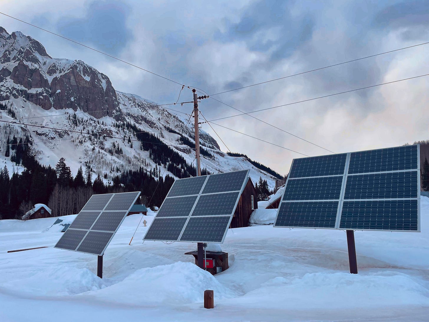 Three solar panel arrays protrude above deep snow near a large mountain under a partly cloudy sky. Three solar panel arrays protrude above deep snow near a large mountain under a partly cloudy sky.