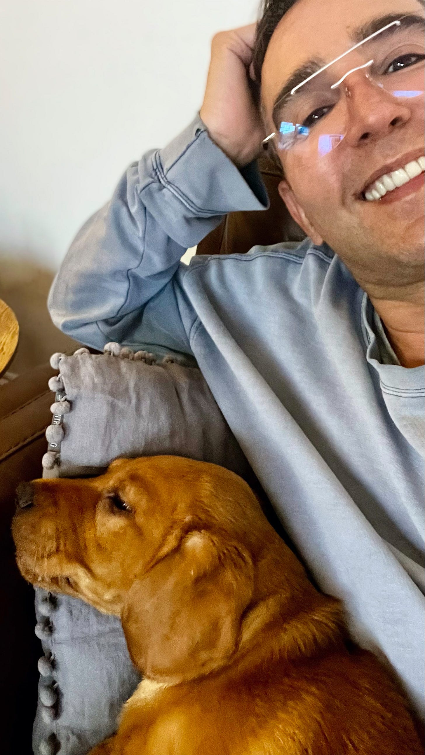 A person with glasses, smiling warmly while reclining on a sofa, resting their head on their hand. Next to them, a brown dog is lying on a cushion, seemingly relaxed.