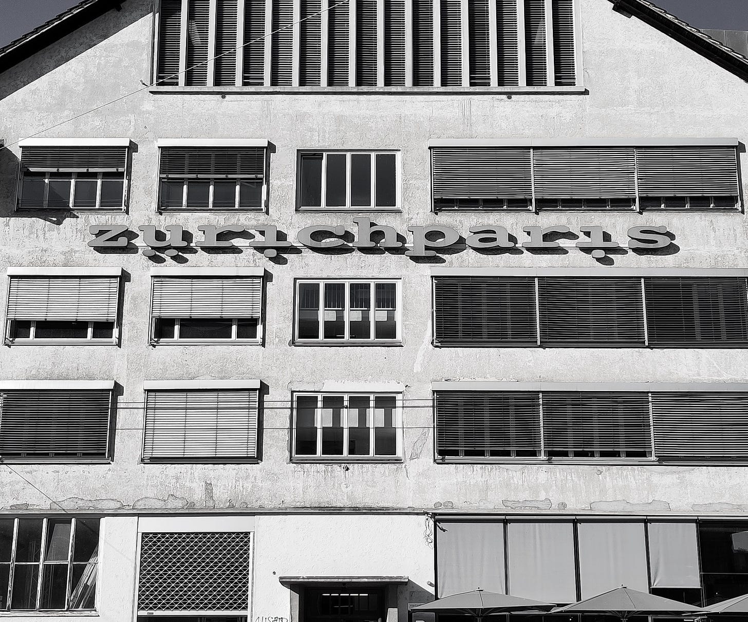 Fotografia in bianco e nero di un palazzo moderno con finestre in parte oscurate da tapparelle e con un'insegna che dice "zürichparis"