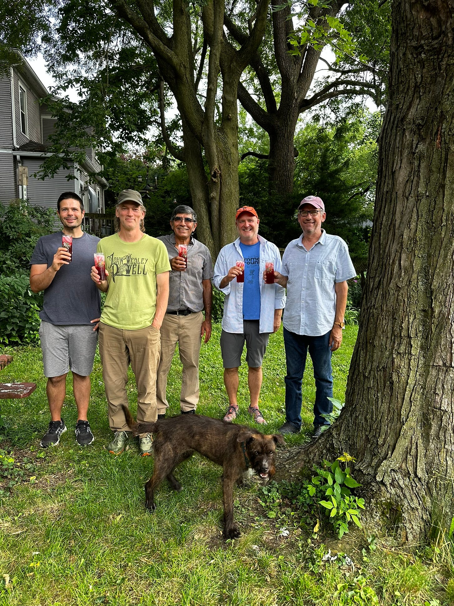 In this image, five men stand smiling under large trees, each holding a drink called a mulberry shrub. The drink is deep red with ice cubes. A dark brown dog stands in the forefront.