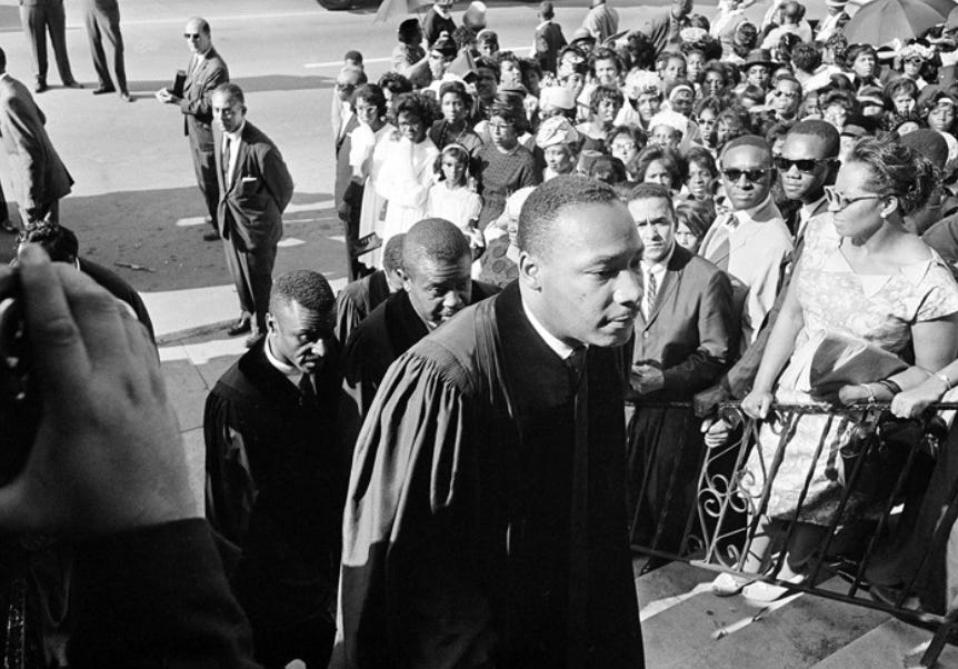 Black and white photo of MLK in Black clerical robes walking up the stairs of the church through a crowd