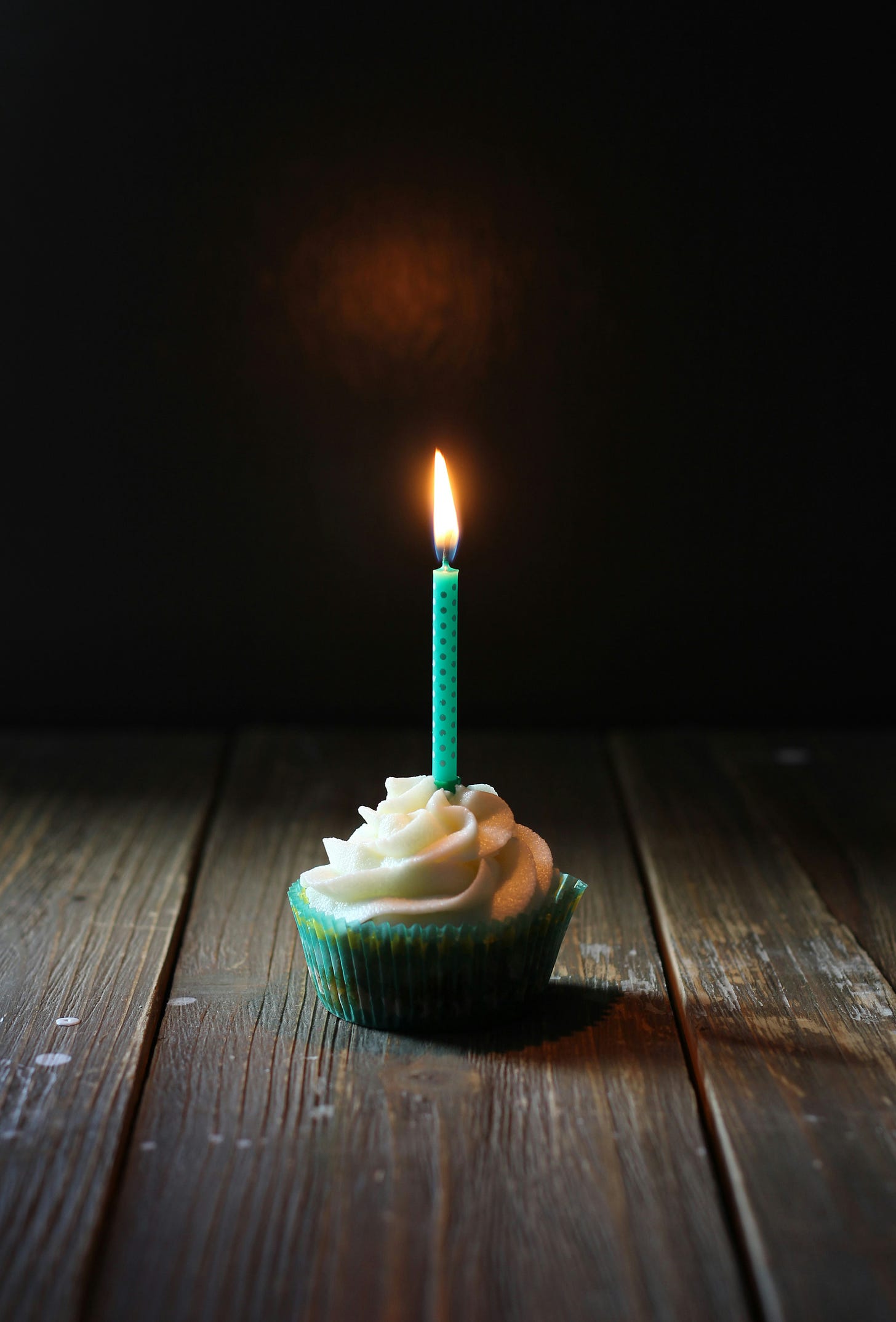 A single cupcake with a mound of curled white icing, in a green wrapper. The cupcake sits on a wooden surface and is illuminated against a dark background. There is one single teal candle in the cupcake and the flame burns merrily. 