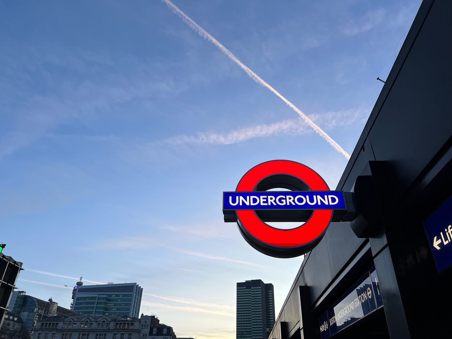 An Underground tube stop sign lit brightly against the blue late afternoon sky with London buildings in the distance
