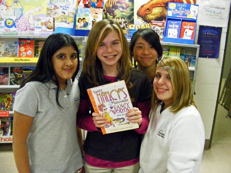 A racially diverse group of four grinning elementary school girls show off a book titled 'Fancy Nancy's Favorite Fancy Words' at a Scholastic book fair. Behind them are wheeled display cases for a range of readers, as well as posters promoting books about dinosaurs, Spongebob, and other 'Fancy Nancy' books, as well as a stand-alone display of the first two books in the 'Diary of a Wimpy Kid' series.