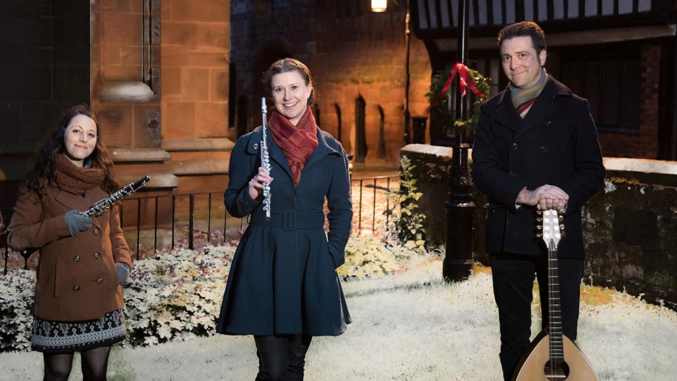 Three members of GreenMatthews stand with their instruments in front of a snowy church. They are warmly dressed to suit the weather.