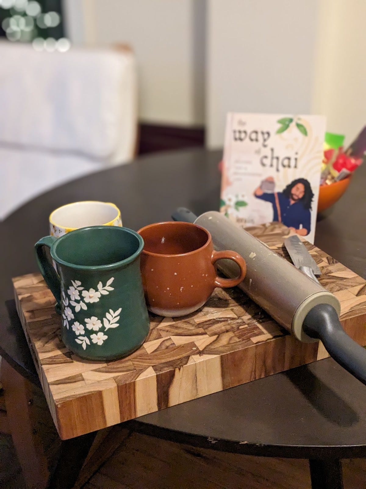 3 mugs sit atop a thick cutting board. There is a green mug with white flowers on it, a smaller and rounder brown mug and a white mug with yellow flowers. Behind is a book title "the way of the chai" with an animation of the author pouring themself a cup of chai