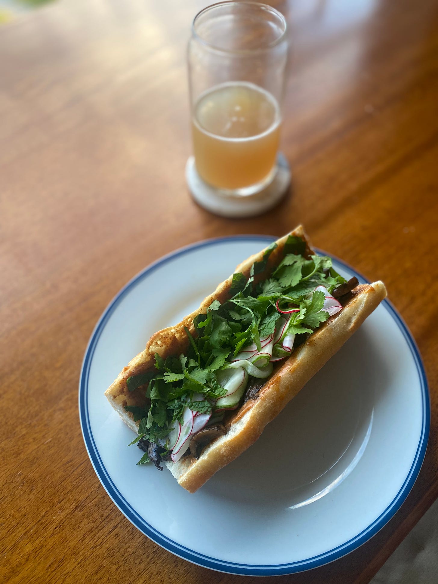 The banh mi described above, viewed from above, with lots of herbs and quick pickles visible. The baguette is open but not fully halved, and you can just see the sauce spilling out a bit at the edge, and some of the mushrooms beneath the vegetables. On a coaster behind the plate is a half-full glass of hazy IPA.