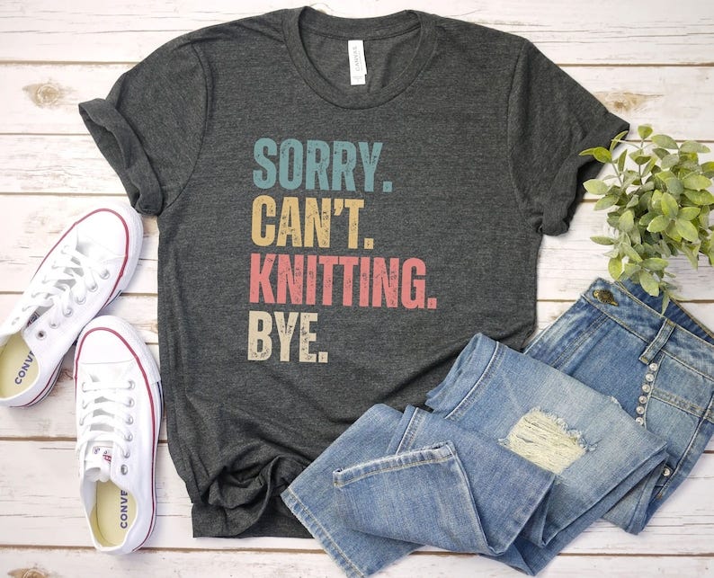 May include: A gray t-shirt with the text 'SORRY. CAN'T. KNITTING. BYE.' in a vintage style font. The text is in various colors, including blue, yellow, red, and orange. The shirt is displayed on a white wooden surface with a pair of white sneakers and blue jeans.