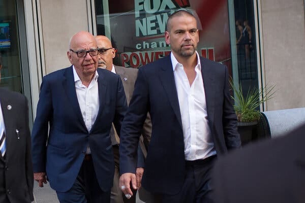 Rupert Murdoch and his son Lachlan Murdoch, both wearing dark suits and white dress shirts, walking with other men. A nearby building’s window has the words “Fox News” on it.