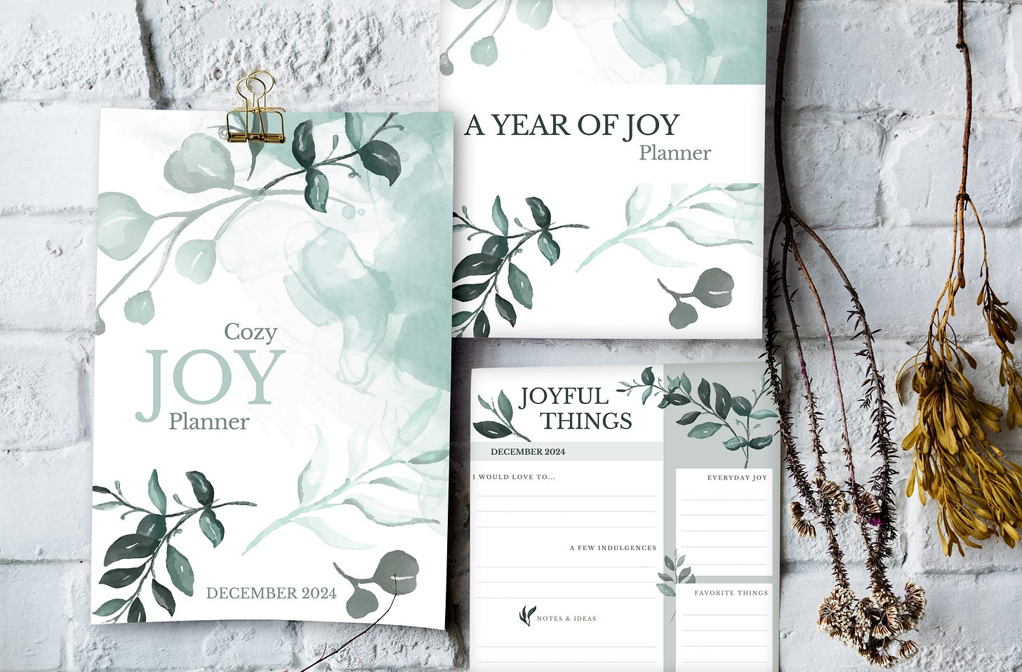 Three pages of the Joy planner hanging on a white brick wall, surrounded by pretty dry twigs