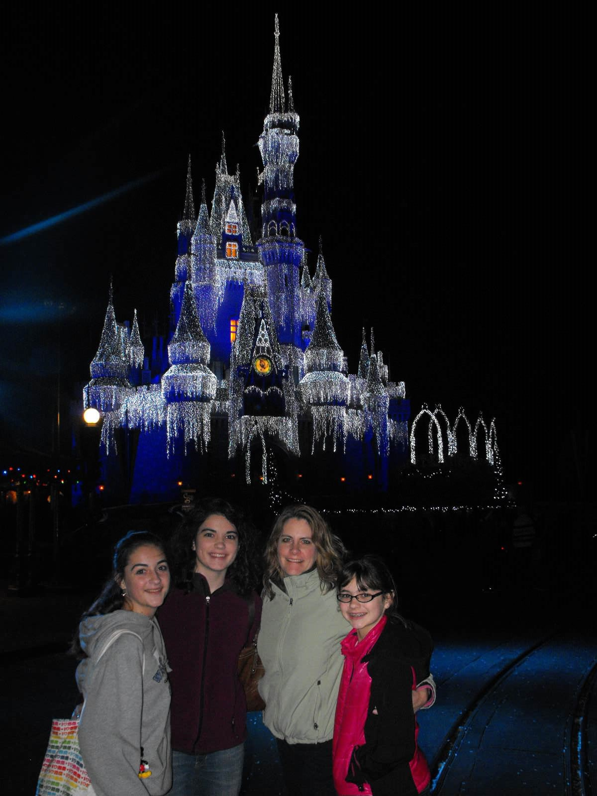 Mom posed with three teen daughters in front of Disney's Cinderella Castle, which is draped in lights that resemble icicles