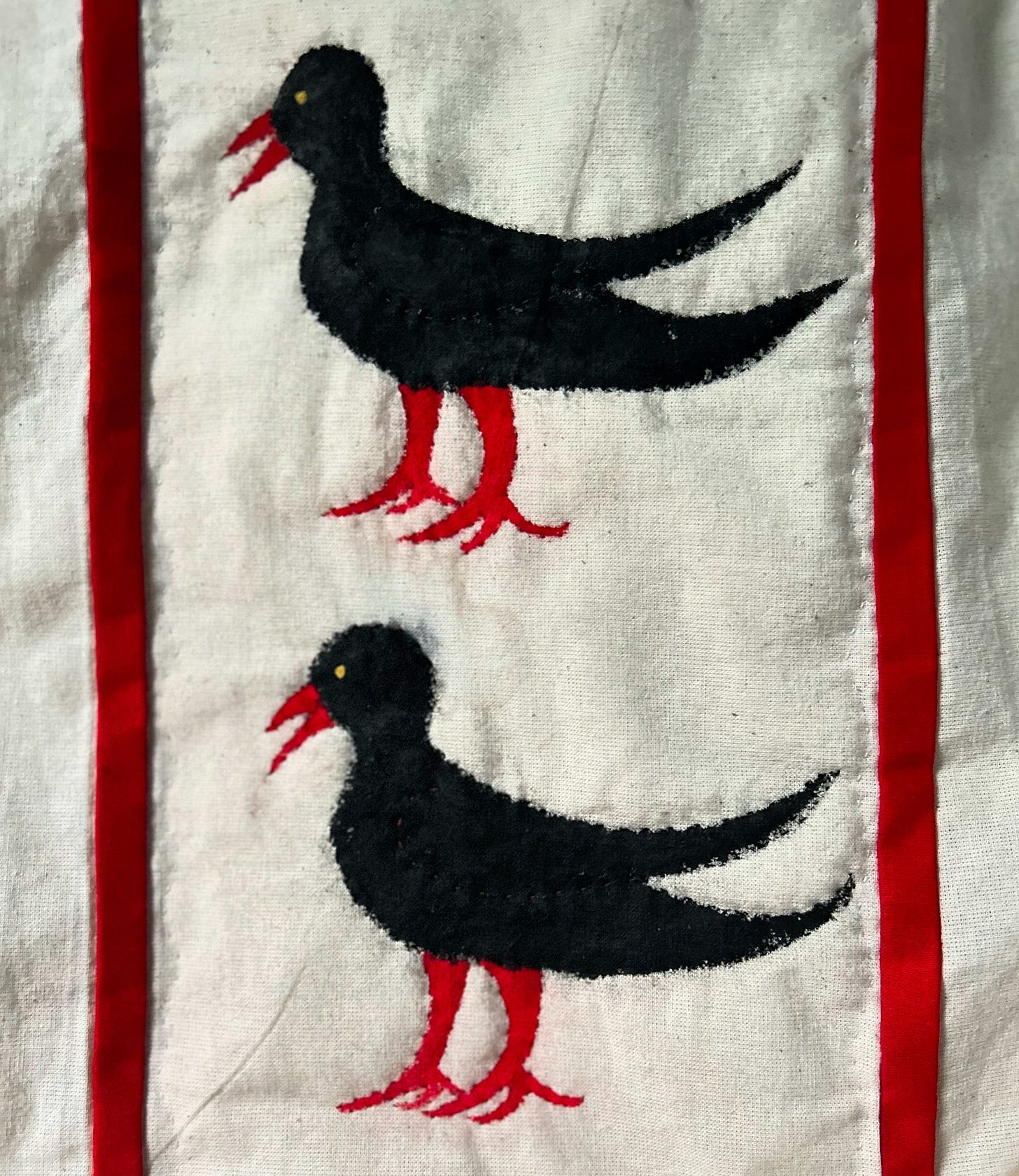 Two black birds with red beaks and legs, painted and stitched onto cream cloth, one above the other,