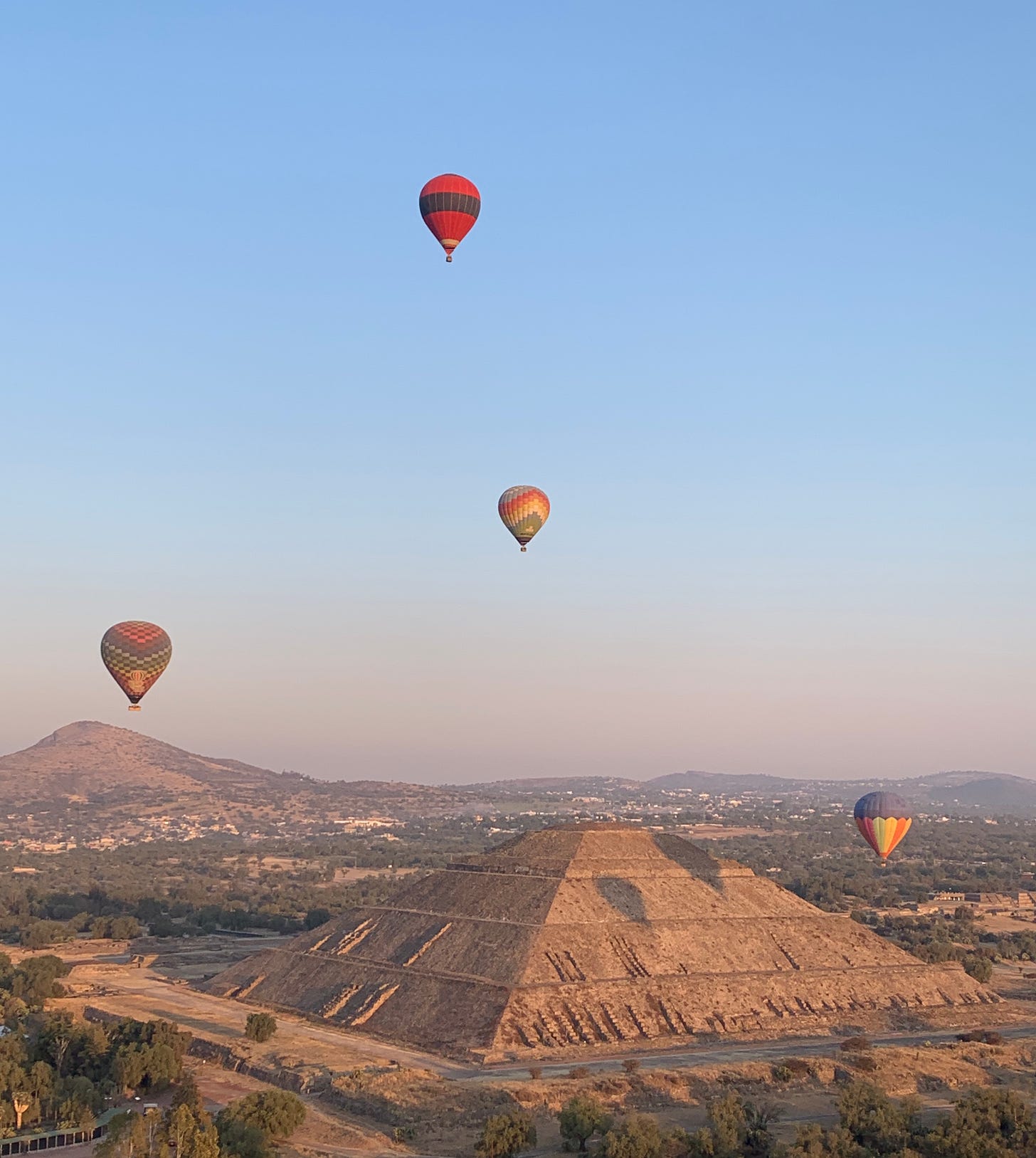 Photo of pale blue early morning sky with four brightly coloured hot air balloons, over two large stone pyramids