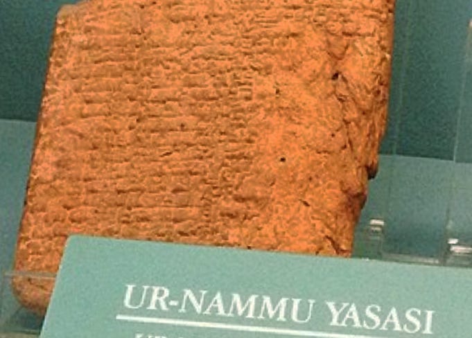 An upright slab of orange clay with hieroglyphic hash marks pressed upon it. Slab is under glass in a museum, and the exhibit sign reads, “Ur Nammu Yasasi,” or Ur Nammu Law Code.