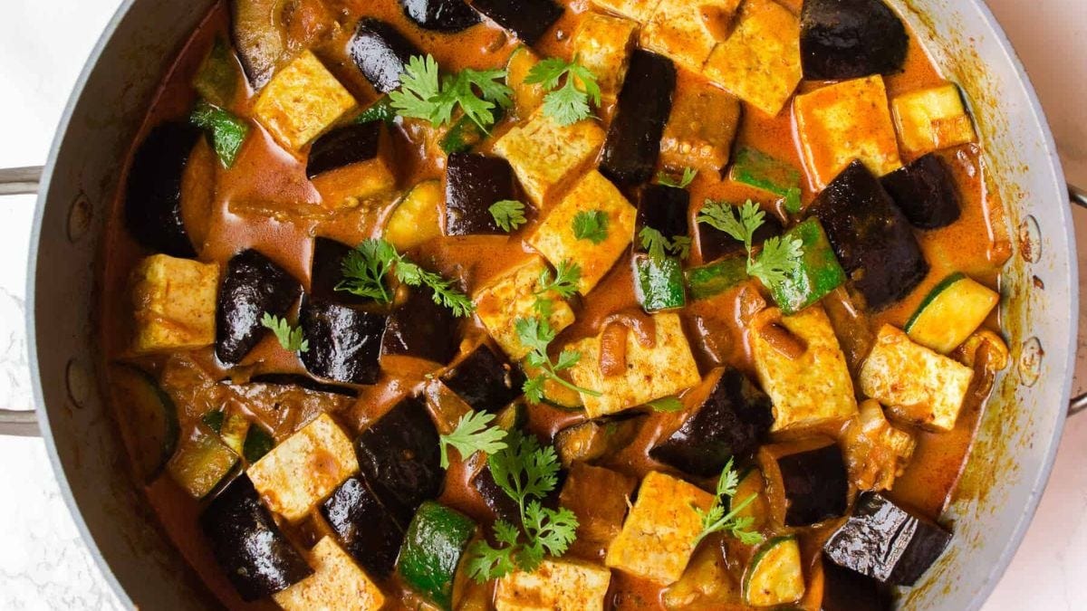 A vibrant pot of tofu and eggplant curry garnished with fresh cilantro. the dish has a rich, reddish-brown sauce and is cooked in a white pot.