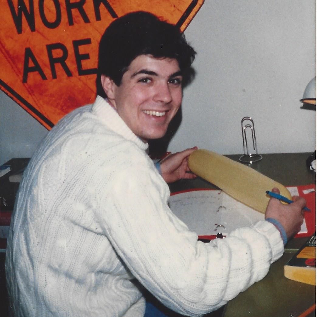 College-aged guy, sitting at a desk doing homework, smiles for the photographer
