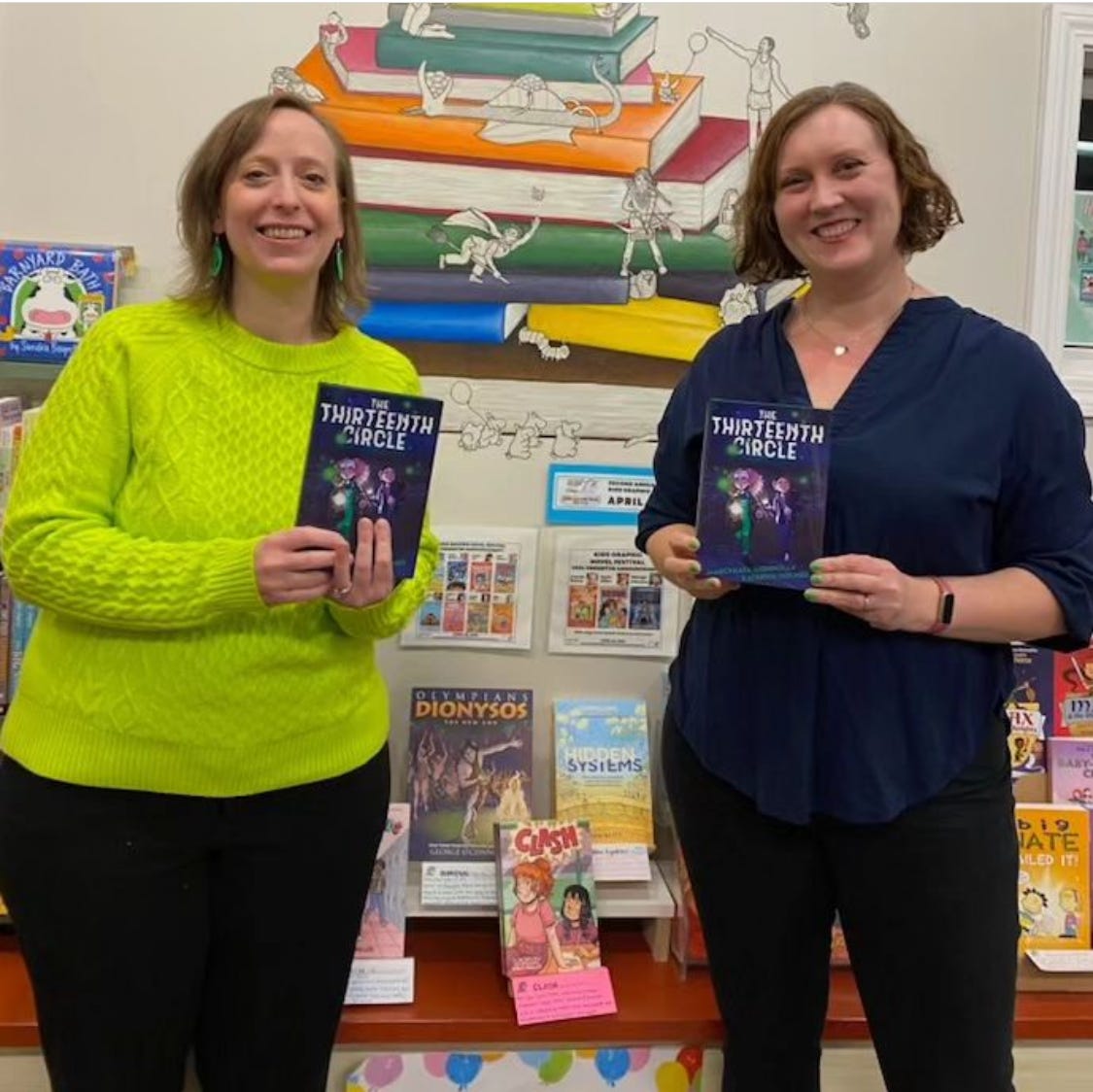 MarcyKate and Kathryn pose with finished copies of The Thirteenth Circle. MarcyKate wears a lime-green sweater and Kathryn wears a navy-blue top, matching their book's cover design.