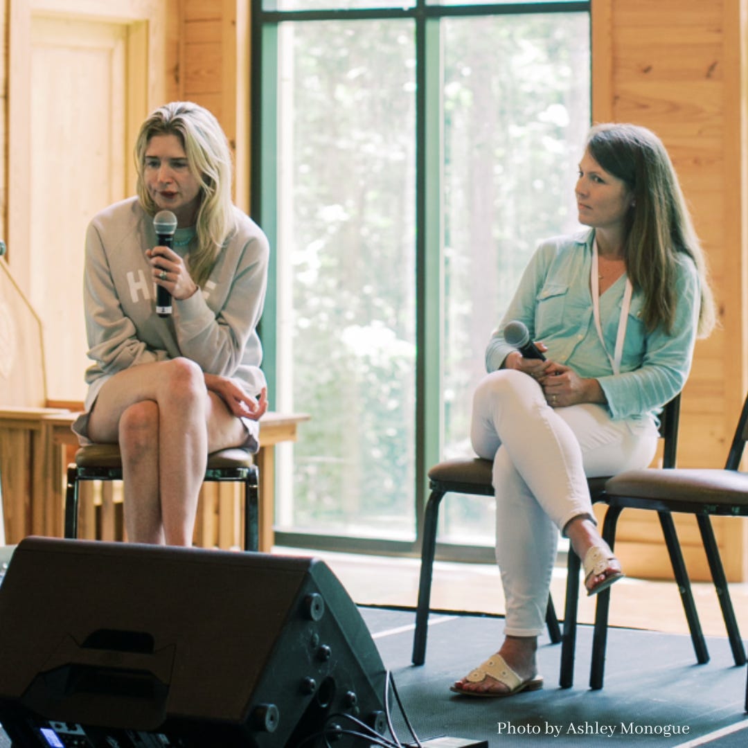 Katherine Wolf and Amy Julia sit on chairs on a stage with large windows behind them