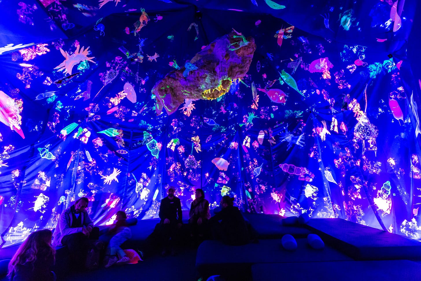 An installation view showing people sitting in a kind of dark blue tent. A fluorescent sea creature hangs from the ceiling. In addition, the fabric of the tent is printed with colourful, fluorescent sea creatures.