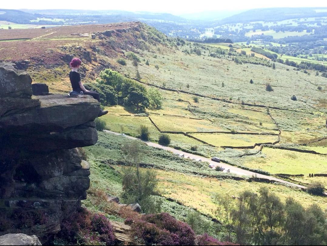 Becky is sitting on a the edge of a rock, looking out at the beautiful view from Curbar Edge in Derbyshire. 