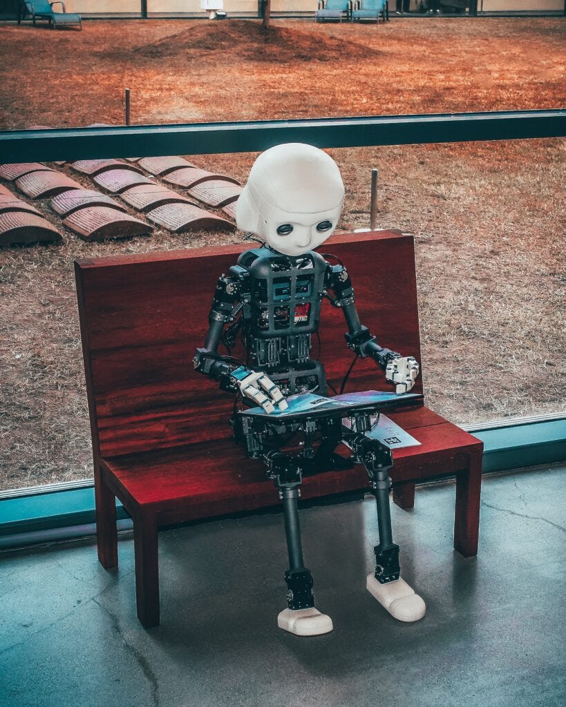 Image shows a robot sitting on a bench with a group of roofing tiles laid out flat behind them to the left. The robot is typing on a keyboard. It's a surreal image.