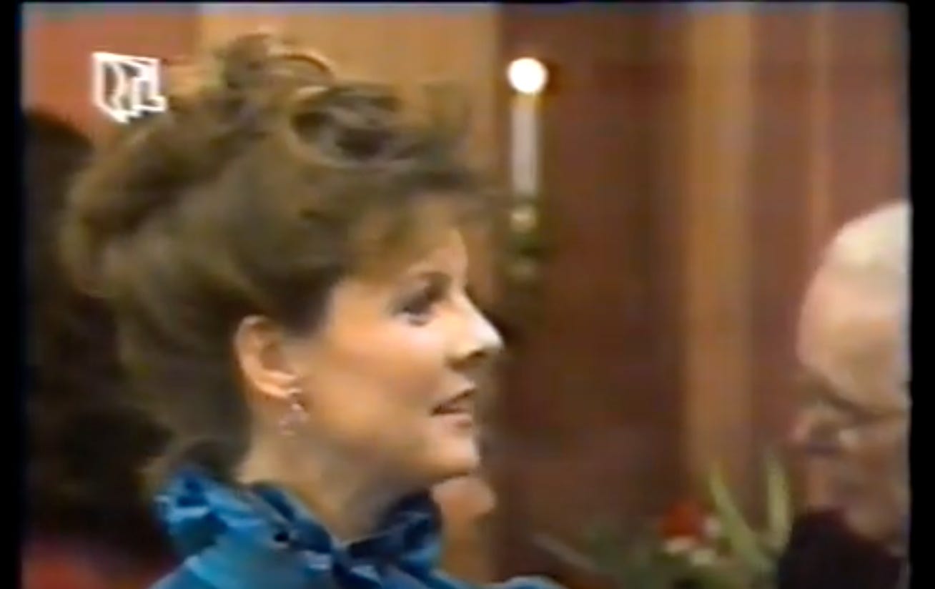 Maureen’s brown hair is pinned up and is wearing a turquoise blouse with a high neck.