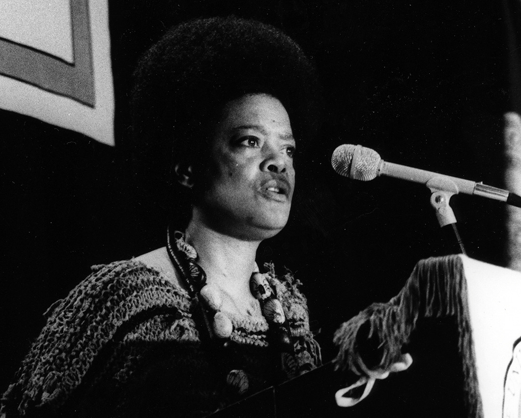 Black and white photo of Toni Cade Bambara looking beyond the camera while wearing a perfectly round afro, a long necklace made of large beads and a knit top. She’s standing at a podium with a mic, lips pursed, mouth speaking into the future.