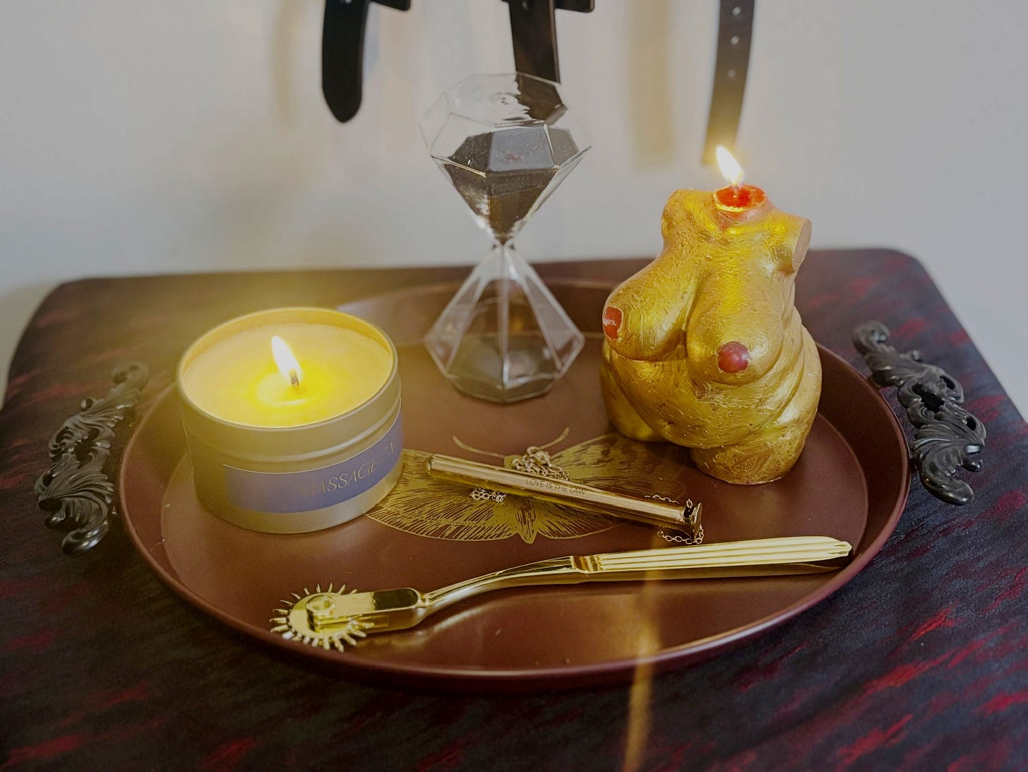 A tray on a bedside table highlighting kinky toys from the Good Ember subscription box. Shown is are two lit candles, a wartenberg pinwheel, an hourglass with black sand and a gold pen shaped Crave Vesper Vibrator