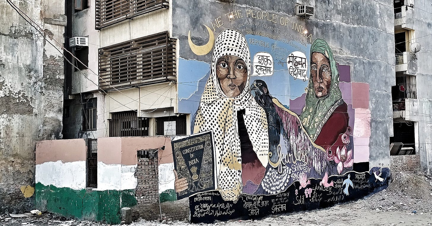 a mural featuring two women, one holding a copy of the Indian constitution. The phrase 'we the people of India' is written above them