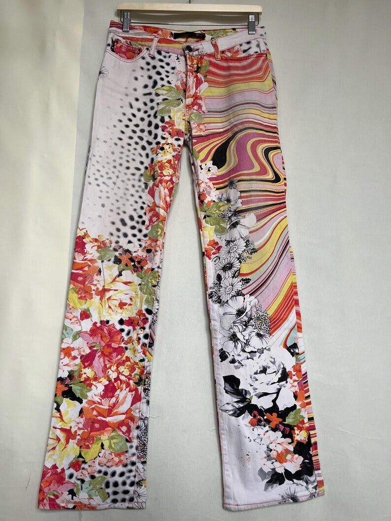 May include: White denim jeans with a colorful floral print. The print includes pink, red, yellow, orange, black, and green flowers. The jeans have a straight leg style.