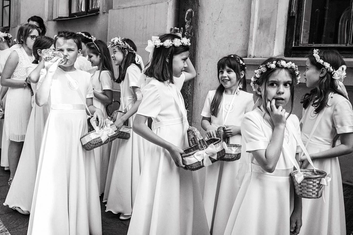 A group of young girls in white dresses and floral crowns stand together outside a building. Some hold small baskets. One girl in the foreground drinks from a bottle while others interact or look around. The image captures a moment of innocence and ceremony, possibly a First Communion or similar religious event.