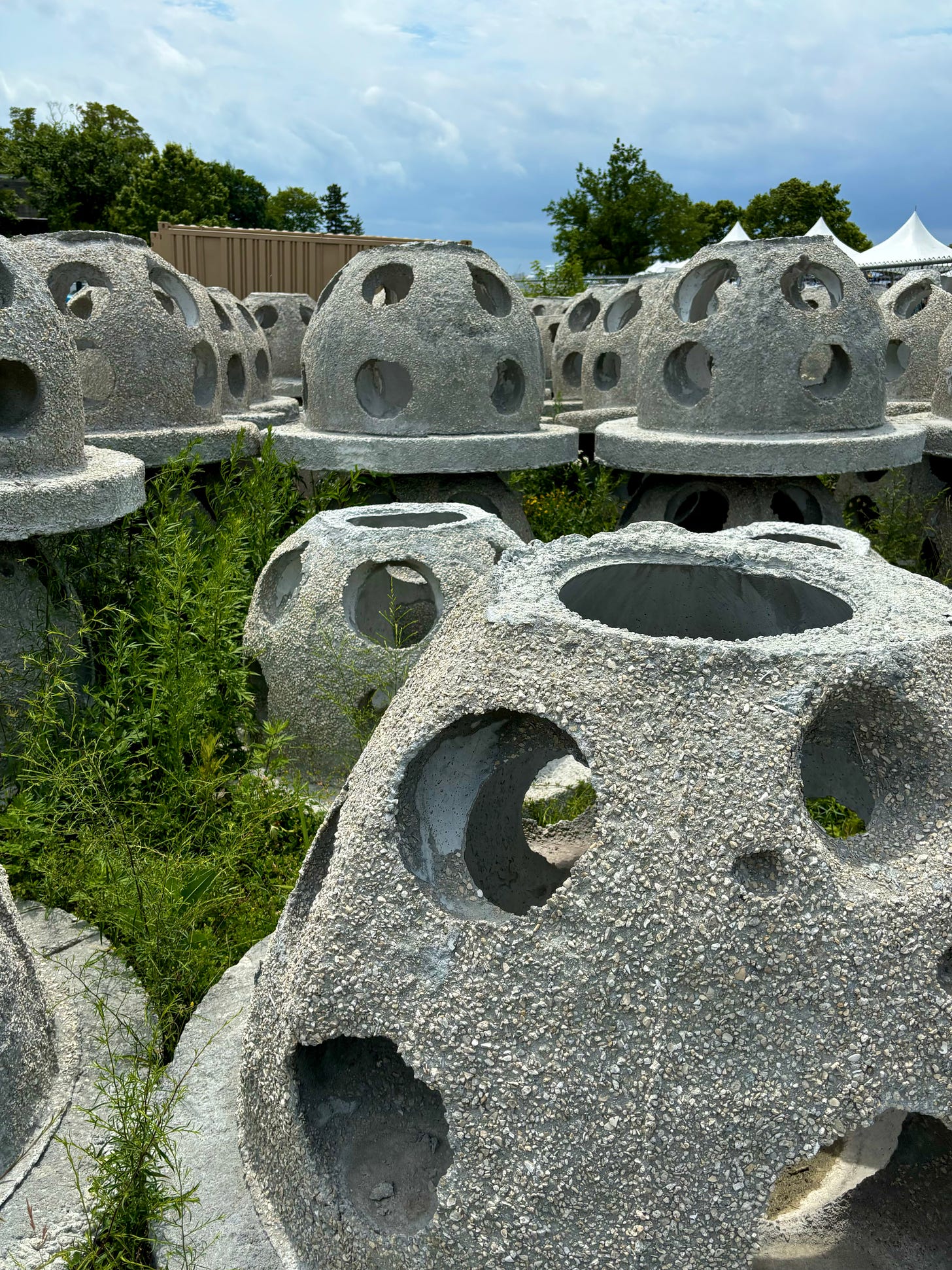 A field of oyster domes stacked two high. They are half-orbs made of concrete with holes throughout like a whiffle ball. There are trees behind and a white tent set-up in the behind them to the right. The tent is for a unrelated party.