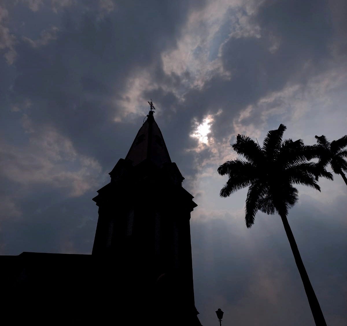 Igreja e palmeira em sombras, com destaque para o céu e o sol tentando sair pelas nuvens