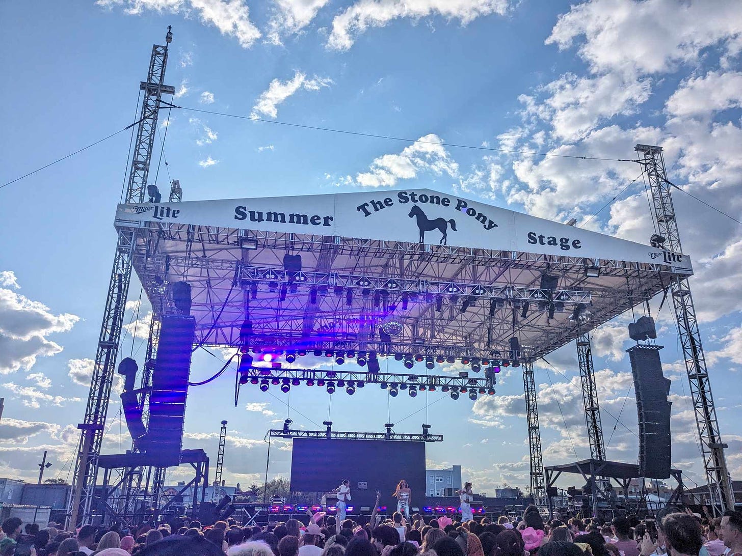 Large stage with a sign that says "The Stone Pony Summer Stage." A white female in silver is standing on the stage with 2 dancers in front of a crowd
