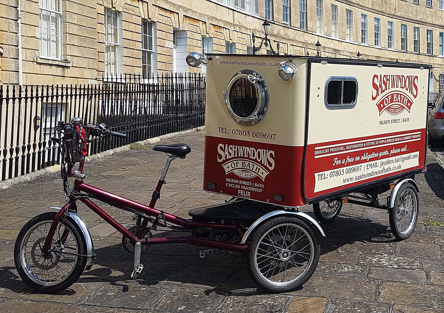 In front of a traditional bath row of town houses, a bicycle with a long covered trailer is parked. On the side of the trailer is an advert for the company