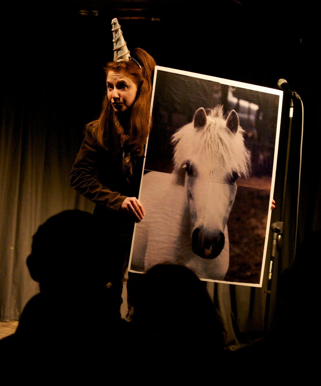 Woman on stage wearing a unicorn horn holding a massive poster of a horse