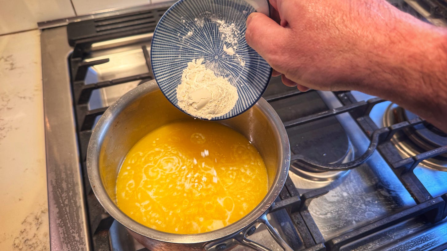 A hand tips the bowl of cornstarch into the pot of melted butter on a stove