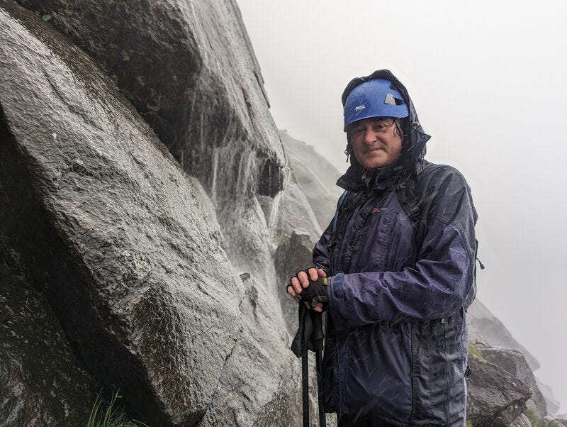 A man in full waterproof and a blue helmet looks grimly at the camera while resting his hands on walking poles. He is standing next to rocks streaming with water. Cloud obscures the background.